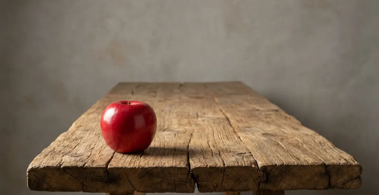 Pomme rouge luisante posée sur une table en bois brut avec grain profond visible, éclairage rasant révélant les textures contrastées