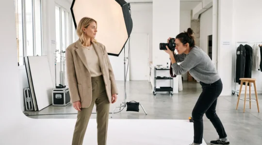 Séance photo mode professionnelle en studio avec mannequin féminin debout et photographe en action cadrant avec appareil reflex, softbox et réflecteurs visibles en arrière-plan sur fond blanc